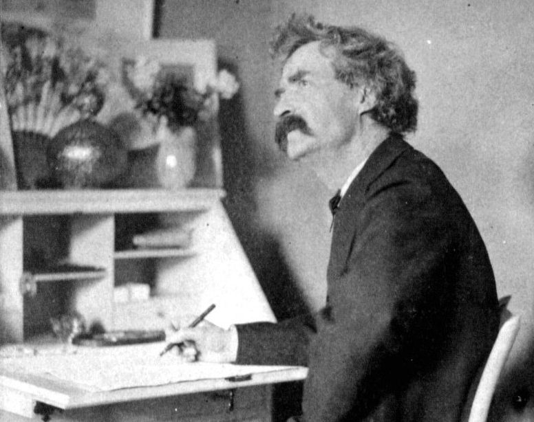 Mark twain sits at his writing desk in this black and white photo with a pen in his hand and is seen looking into space 