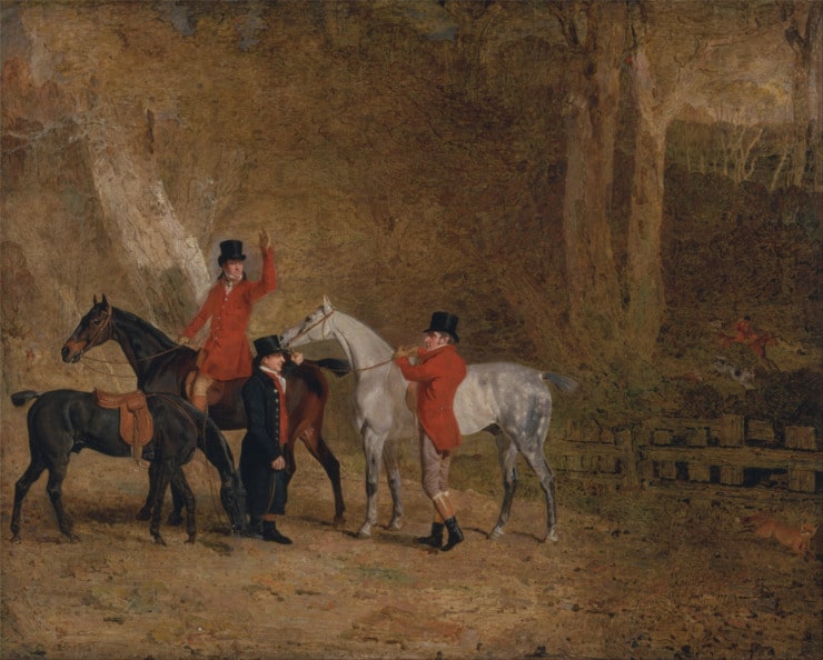 a fox hunter wearing a red long coat rides out of the woods with a partner and two horses.