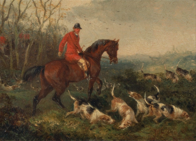 a fox hunter wearing a red long coat rides out of the woods with several hound dogs.
